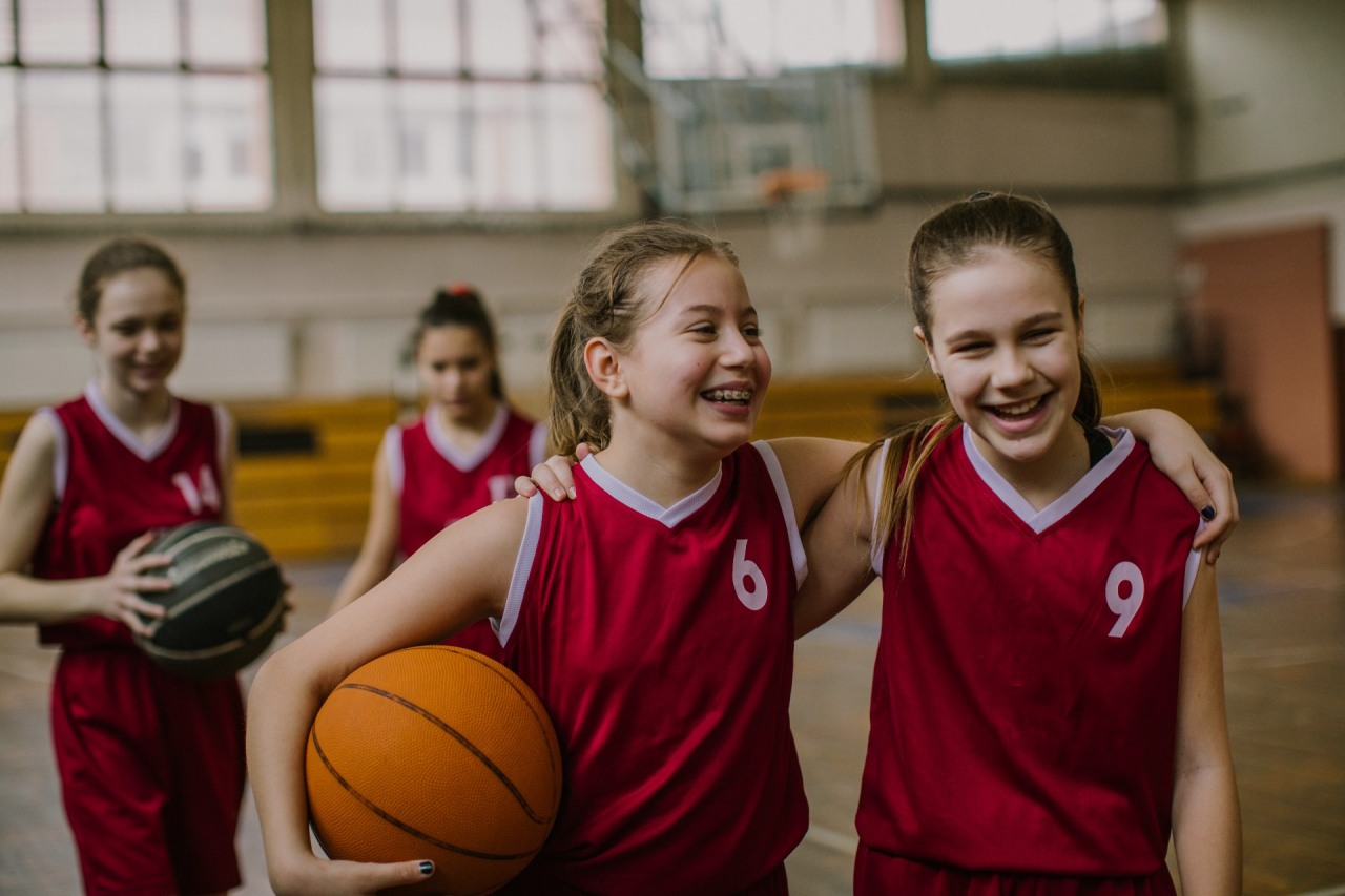 Fundacja OLREN. Dwie dziewczynki w strojach koszykarskich grają w koszykówkę na szkolnej hali.