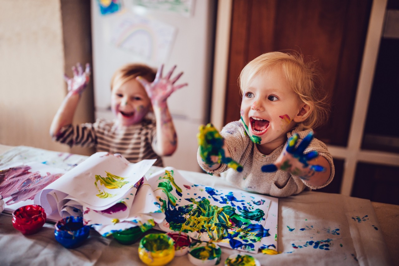 Dwie małe dziewczynki malują obraz palcami. Dłonie oraz twarze są pokryte plamami farby.