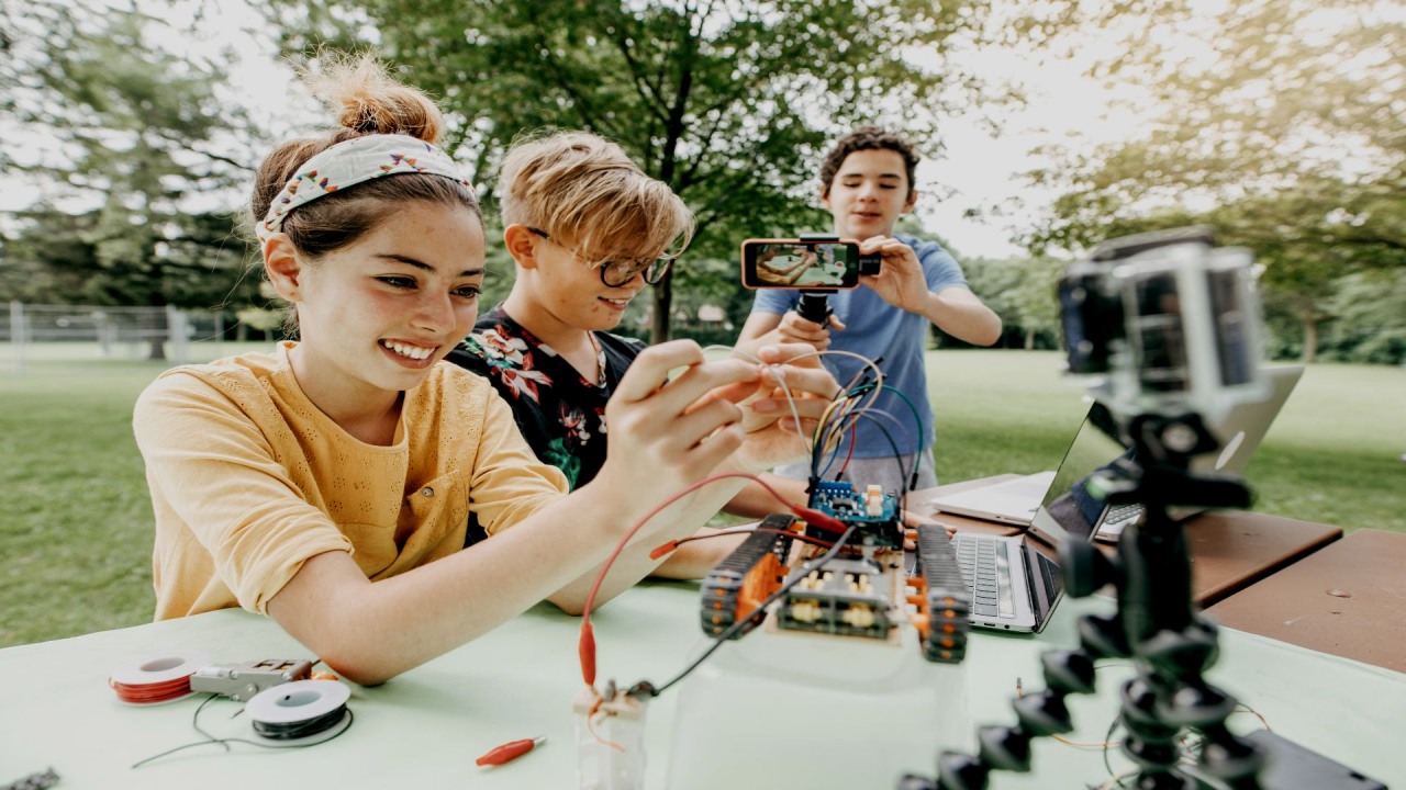 Fundacja ORLEN. Dwoje dzieci konfiguruje robota na świeżym powietrzu. Trzecie dziecko w tle nagrywa całą scenę smartfonem